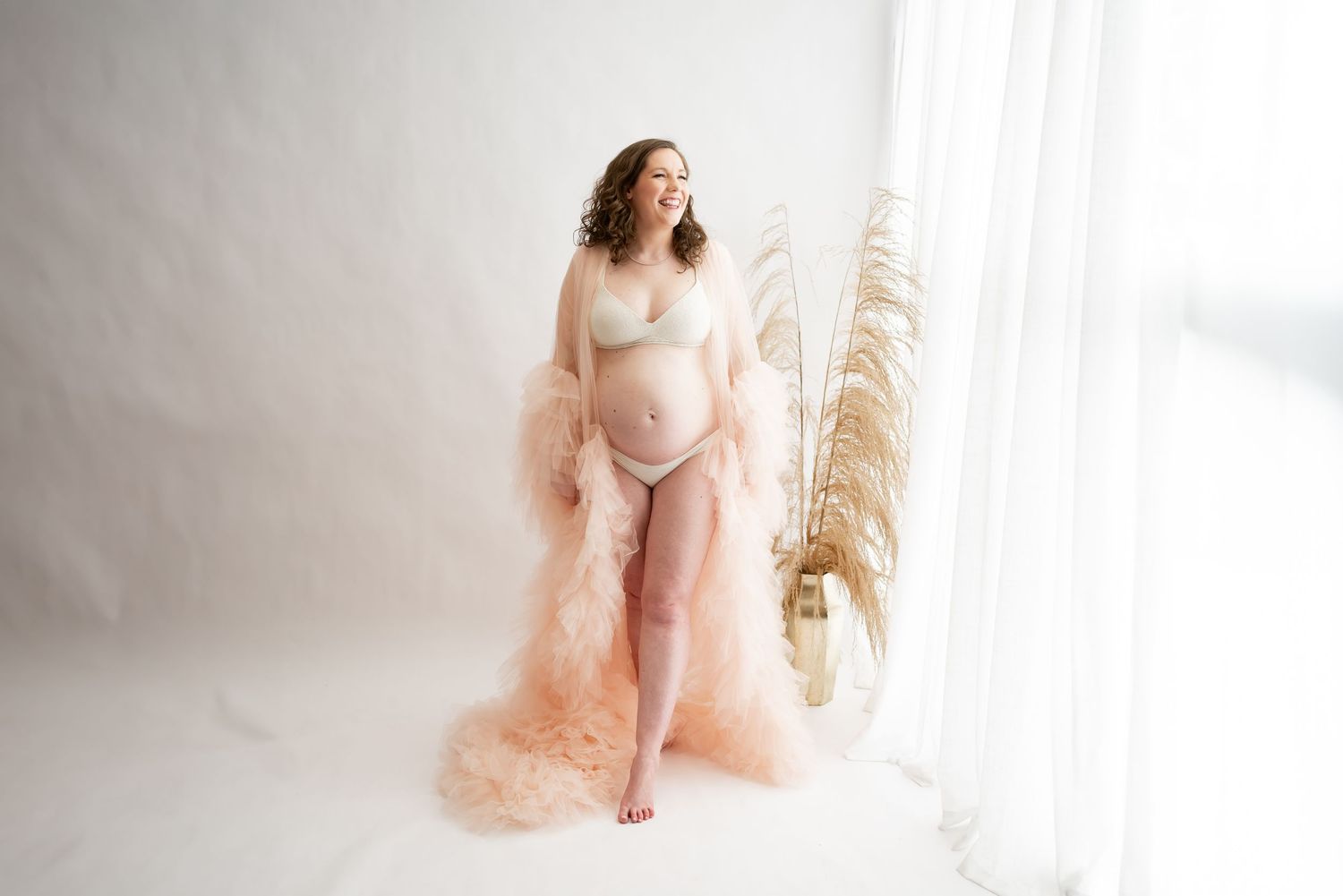 Ethereal figure in flowing champagne tulle gown poses gracefully against white backdrop with pampas grass decoration.
