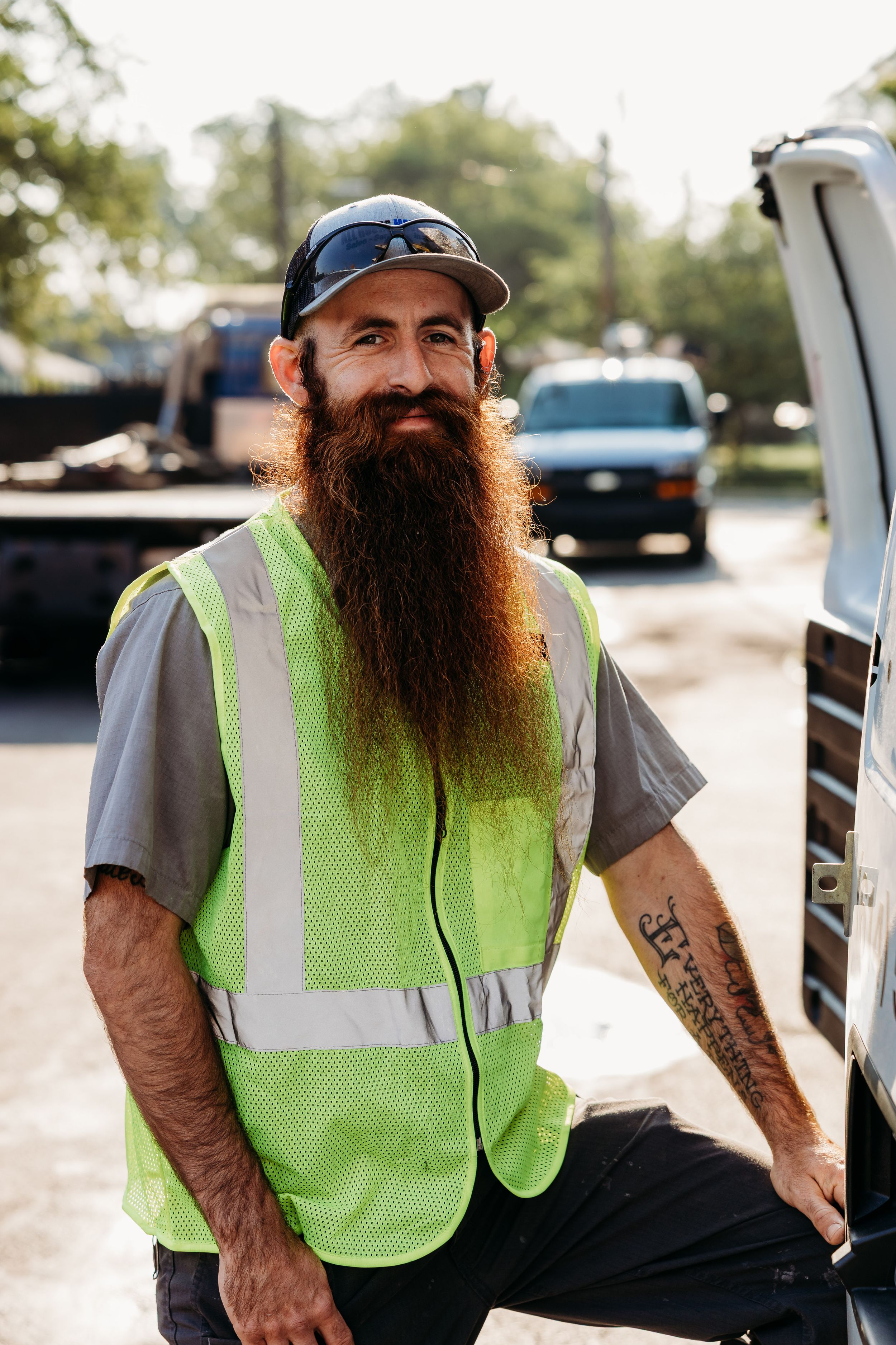 Worker in high visibility safety vest and baseball cap with long beard standing next to vehicle outdoors.