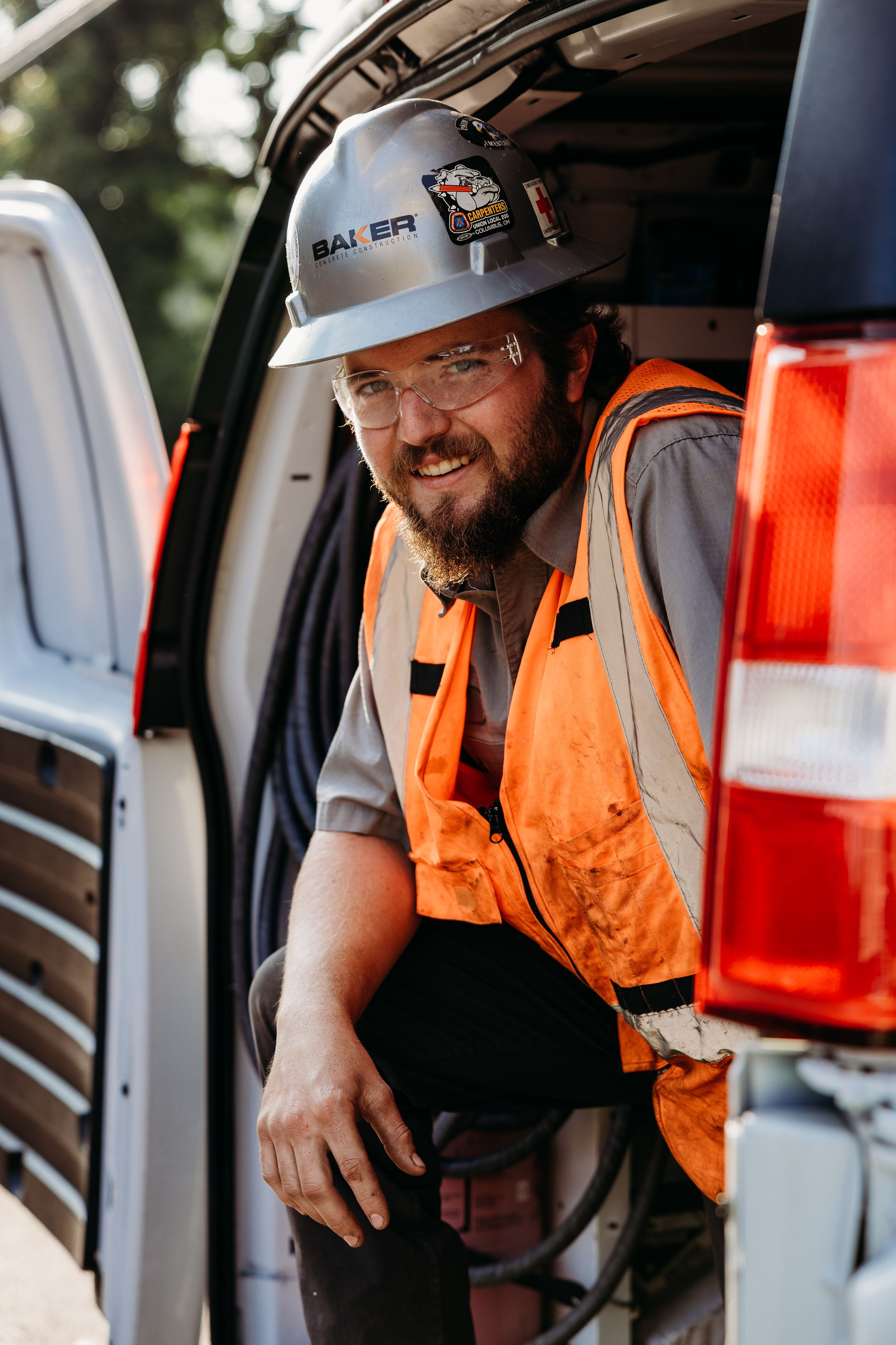 Construction worker in orange safety vest and hard hat sits in work vehicle with a friendly expression.