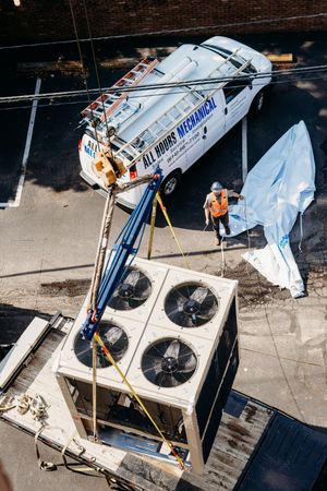 An aerial view of a crane lifting HVAC equipment onto a building with workers and a service vehicle on the rooftop.