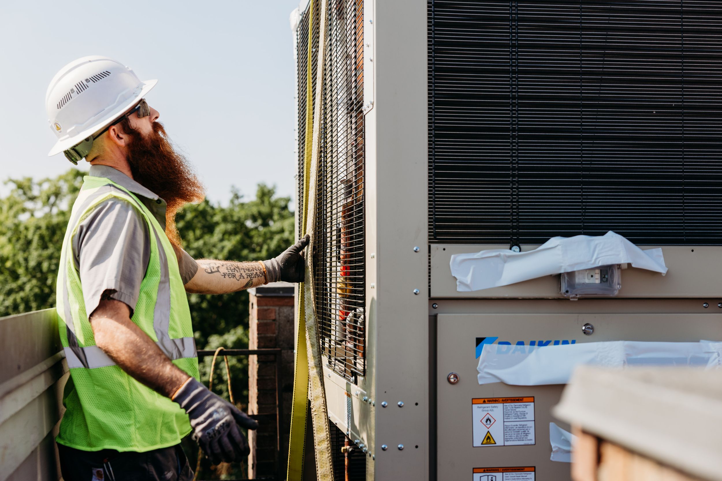 HVAC technician in safety vest and hardhat performs maintenance on commercial air conditioning unit outdoors.
