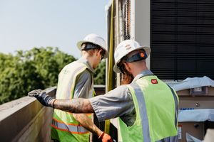 Construction workers in safety gear inspect solar panels on a building rooftop during installation work.