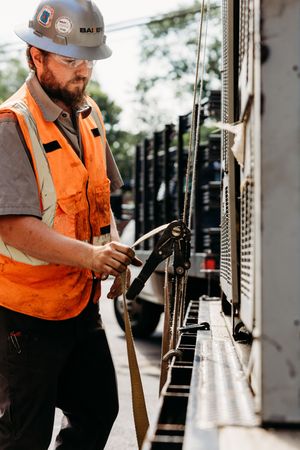 A railroad worker in orange safety vest and hard hat performs maintenance on train equipment.