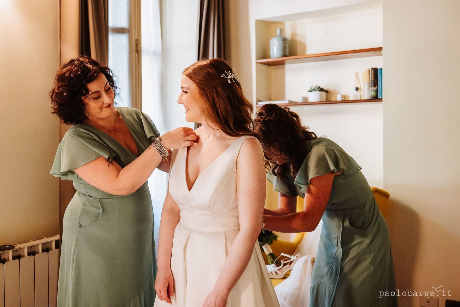 Two women share an intimate moment in a bright modern room with white walls and wooden shelf accents.