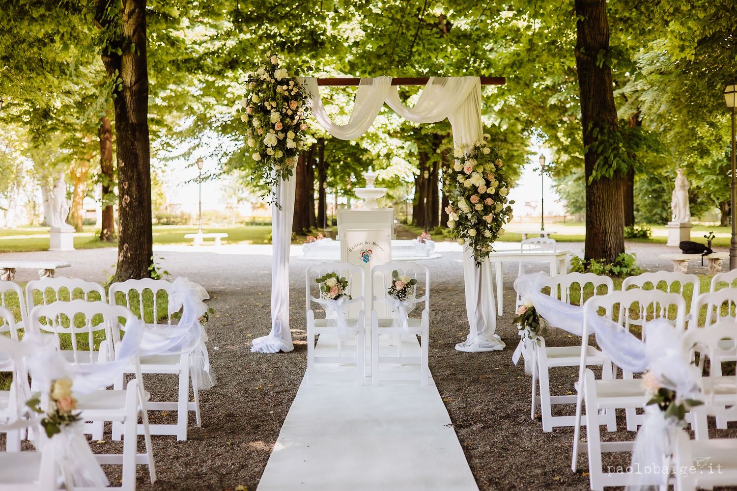 White chairs line a floral-decorated aisle path in an outdoor garden wedding ceremony setup under shaded trees.