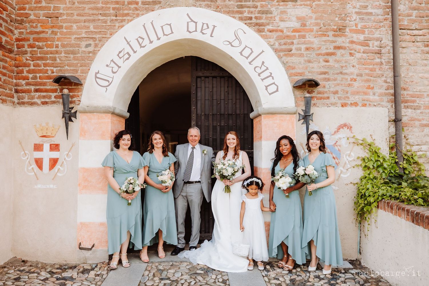 A wedding party in light blue dresses poses beneath a stone archway entrance at Castello del Solaro.