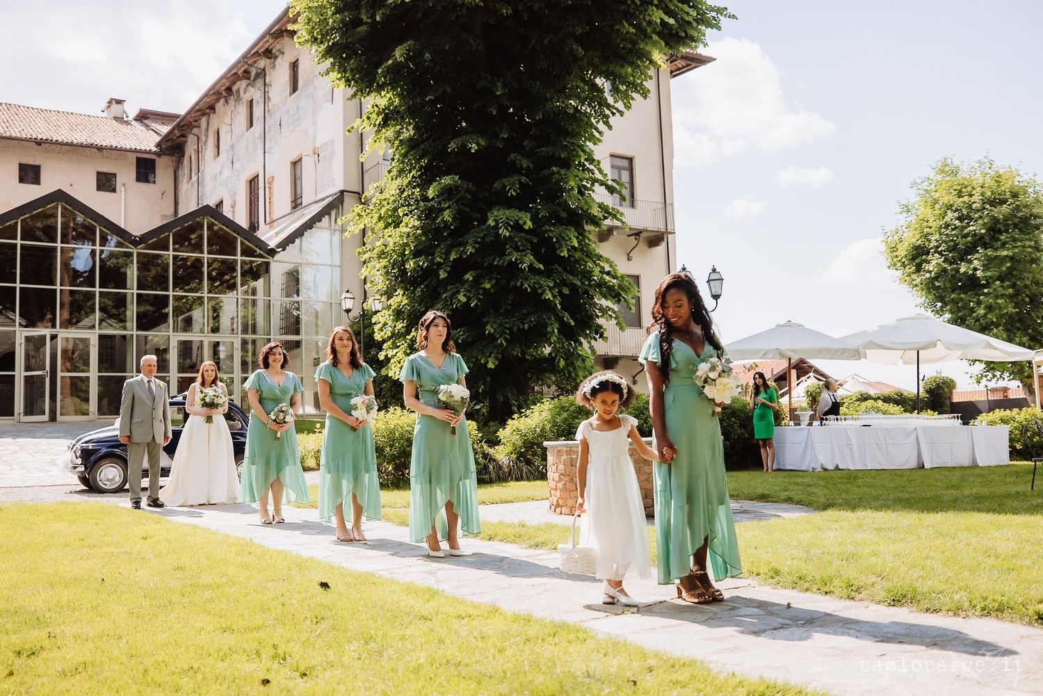 Wedding party wearing mint green dresses poses joyfully on a grassy hillside on a sunny summer day.