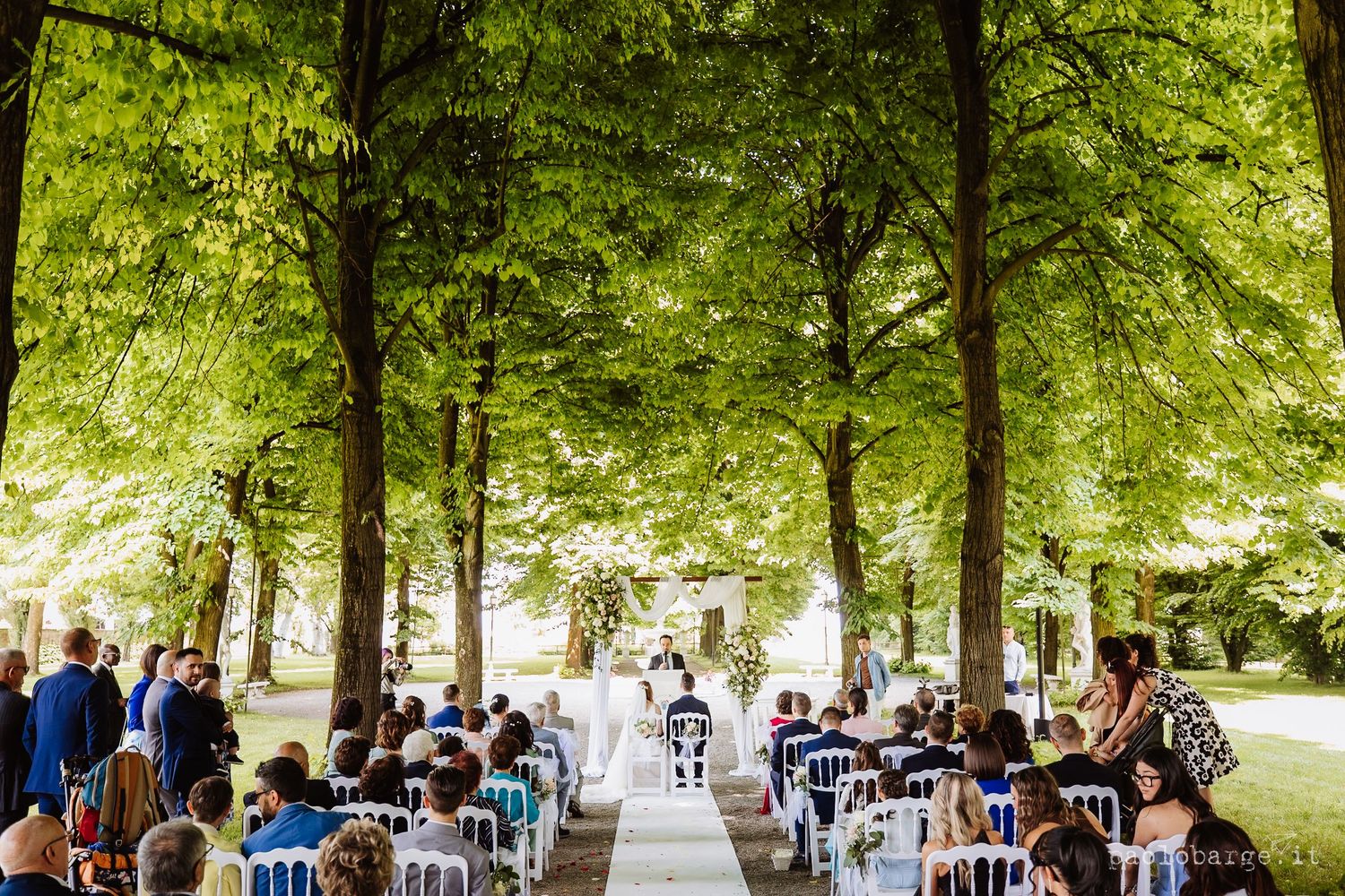 A rustic outdoor wedding ceremony set under tall shady trees with rows of white chairs and natural decor.