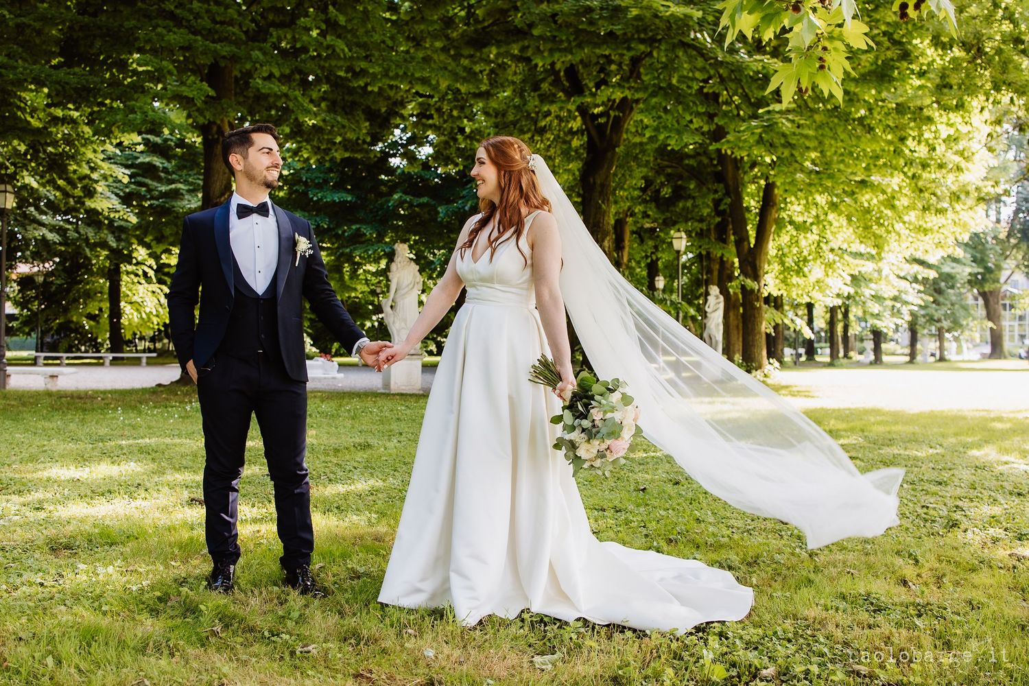 Wedding couple walks together through sunlit park surrounded by lush green trees on sunny summer afternoon.