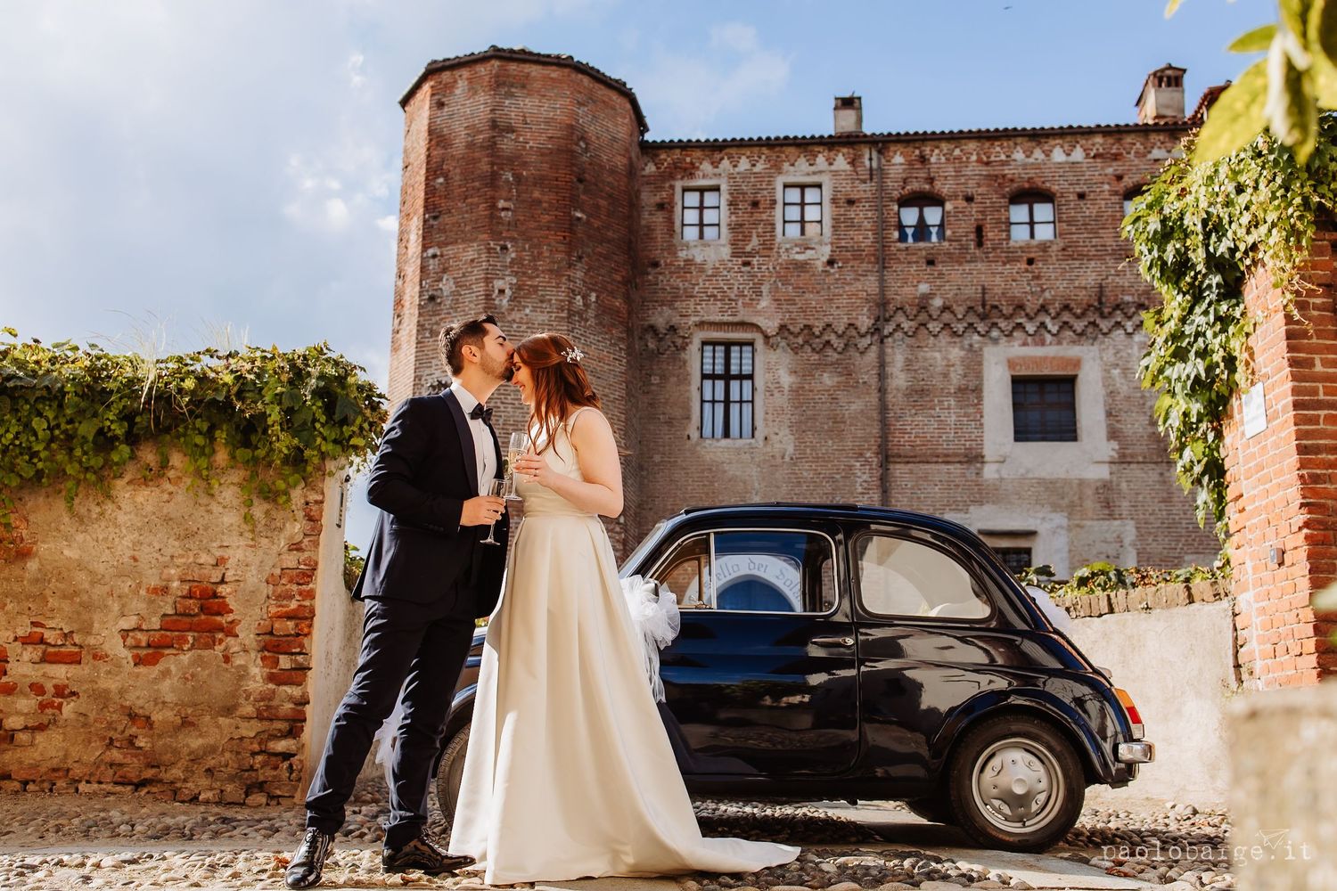 A romantic outdoor wedding scene with a classic vintage car and rustic brick architecture in warm sunlight.