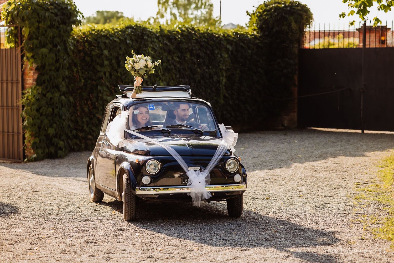 Classic vintage car sitting on a gravel driveway with green bushes and trees in the background.