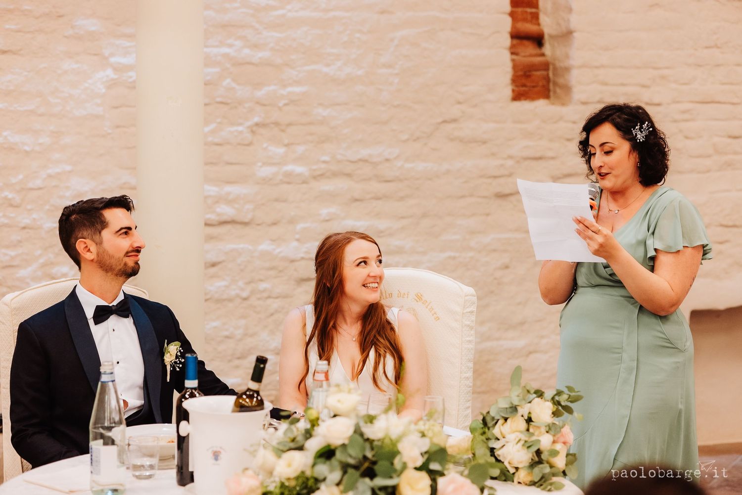 A bridesmaid in a mint green dress reads a speech during a wedding reception while others at the table smile and listen.