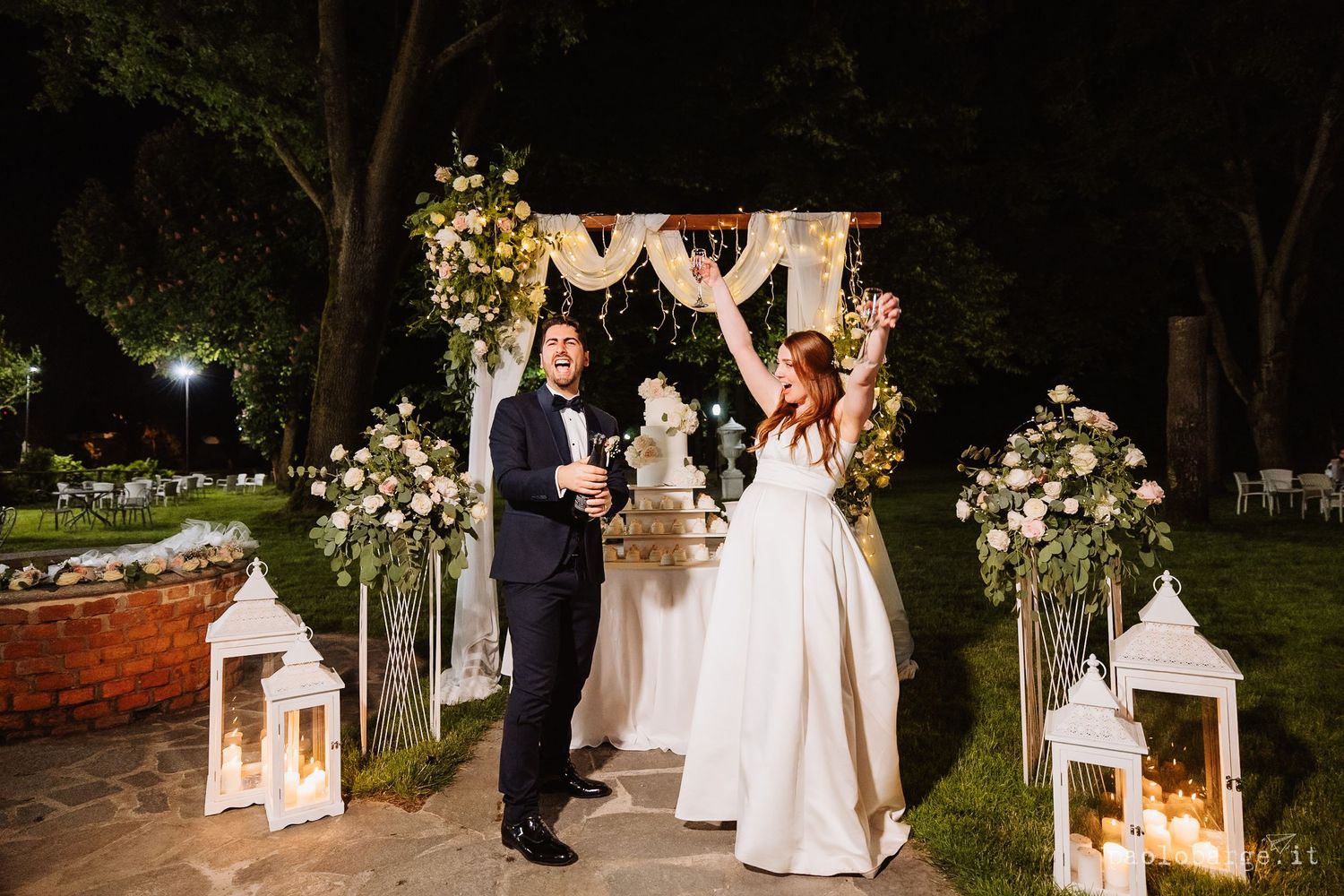 A nighttime outdoor wedding ceremony with illuminated lanterns and floral decorations along a garden path.