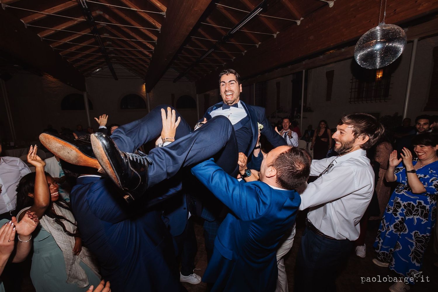 Wedding reception attendees in blue suits lift and carry someone across a wooden beamed venue with a disco ball.