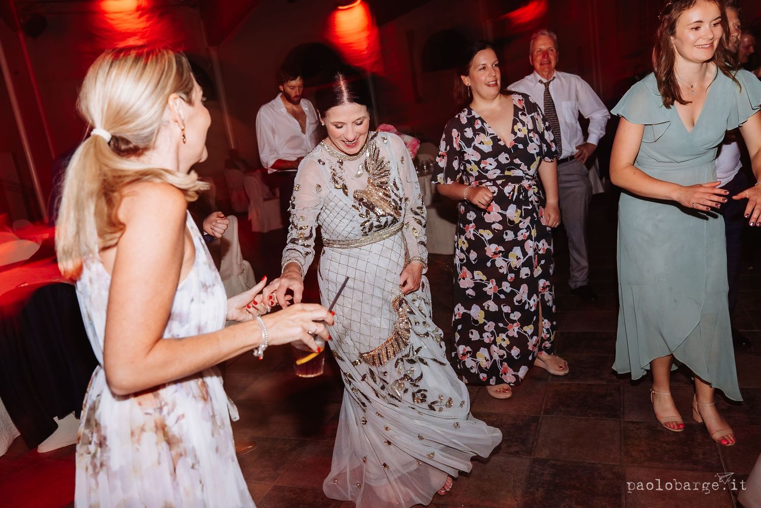 People dancing and celebrating at a wedding reception with red lighting and bright formal dresses on dance floor.