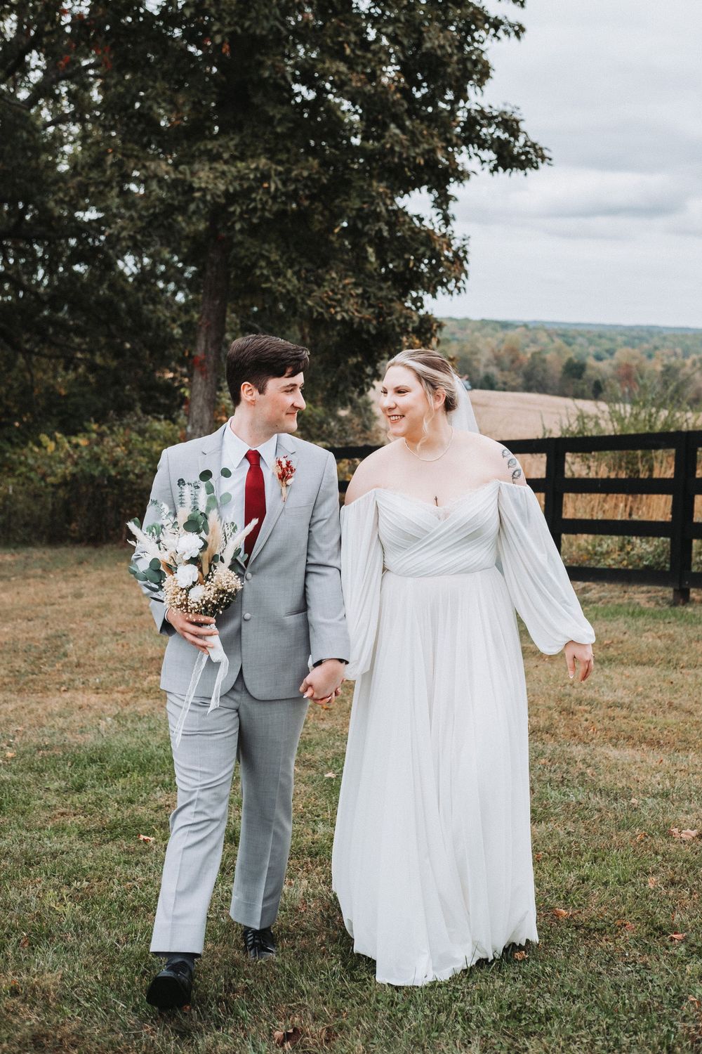 Couple walking hand in hand through scenic field with rustic fence in background.