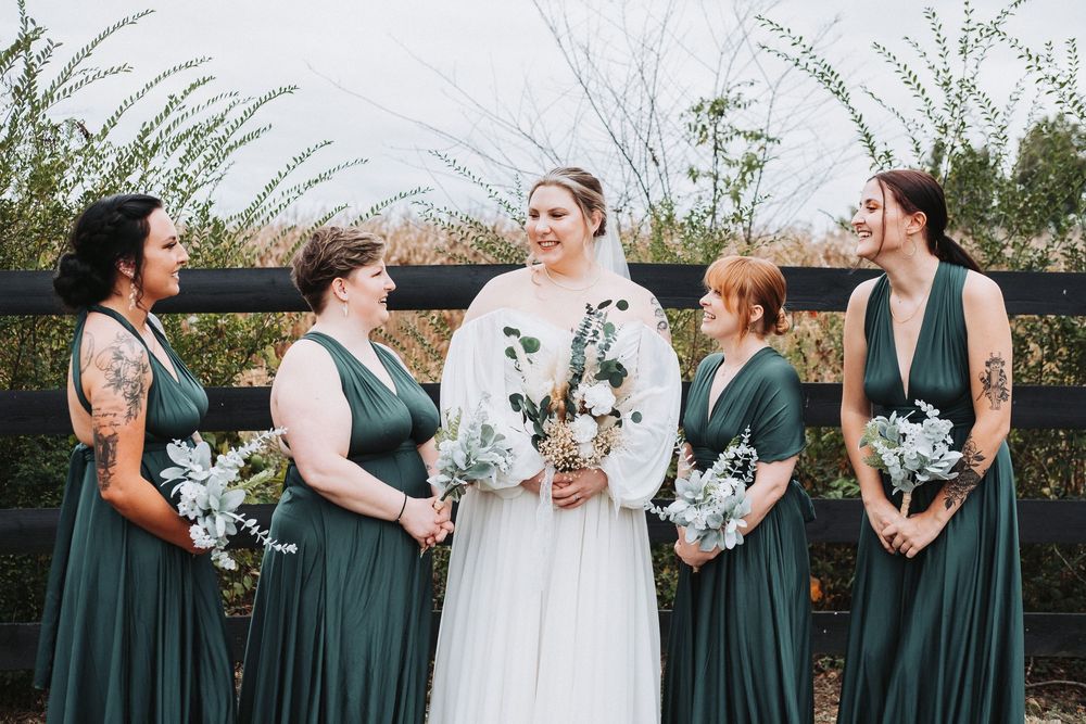 Bride in white dress stands with bridesmaids wearing emerald green gowns holding blue and white floral bouquets outdoors.