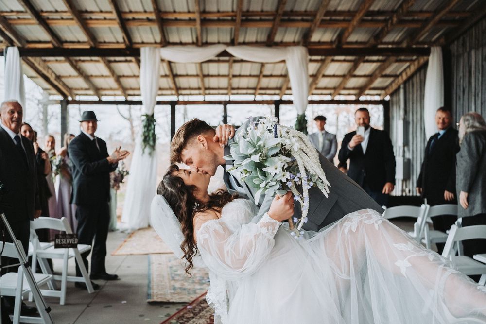 A couple celebrates their first kiss during indoor wedding ceremony in rustic venue with exposed beams and white chairs.
