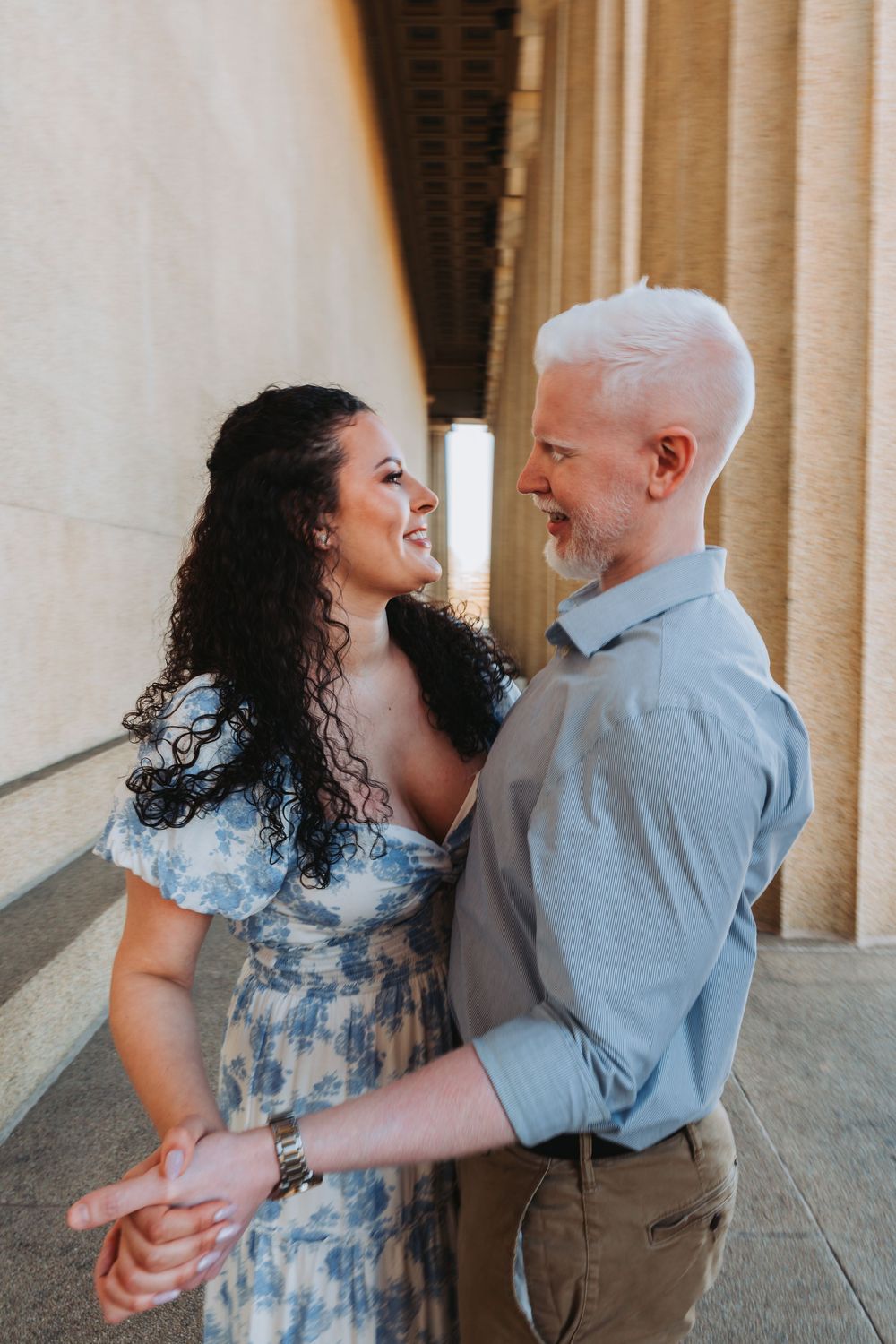 A couple in romantic embrace dance together in an elegant corridor with columns as sunlight streams in from behind.