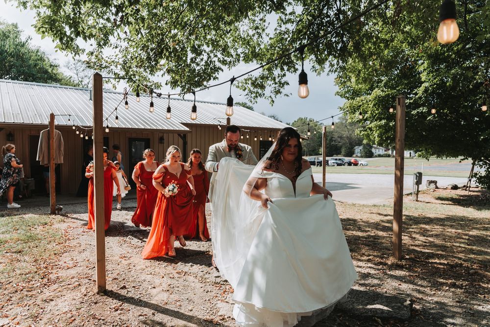 A bride in a white dress twirls outdoors while bridesmaids in orange dresses watch under string lights and trees.