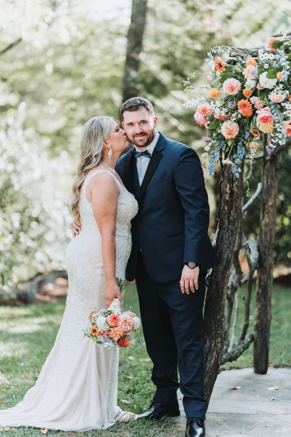 A wedding couple stands under a lush floral arch with peach and white blooms in an outdoor garden setting.