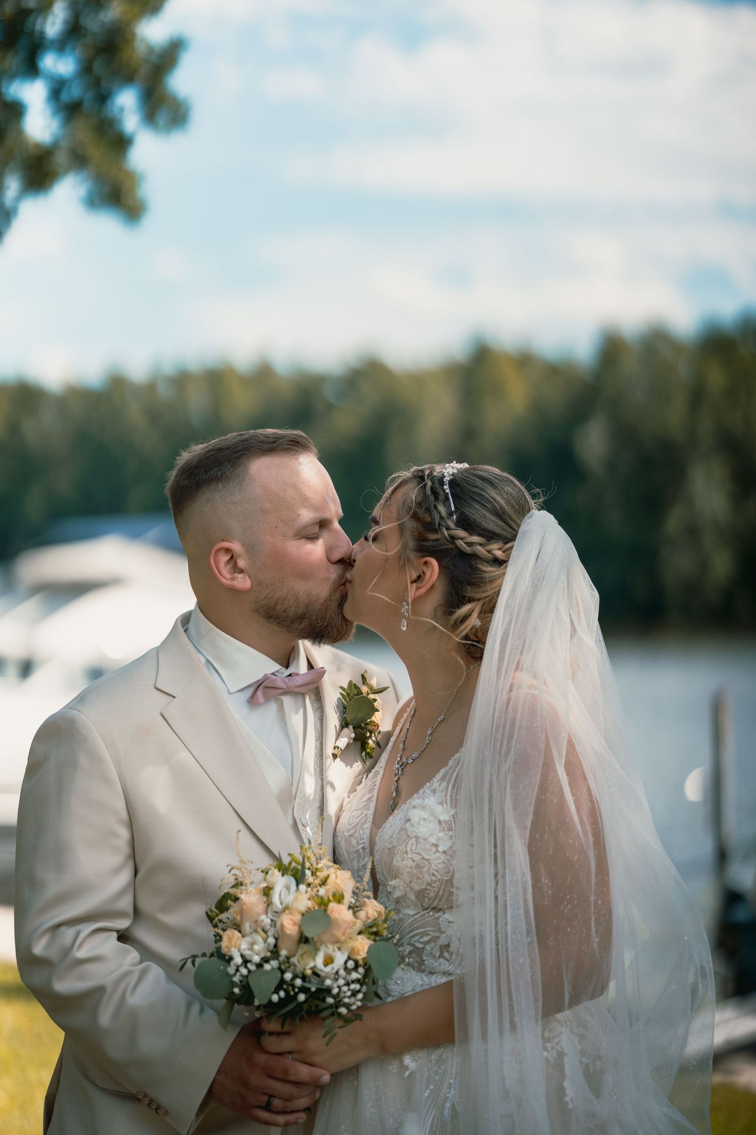 Ein Paar in Hochzeitskleidung teilt einen romantischen Kuss am See, die Braut hält einen Pfirsich- und Cremestrauß.
