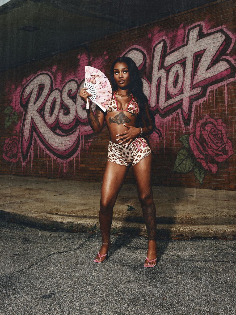 Woman in floral bikini set holding a fan poses in front of a pink rose graffiti mural outdoors.