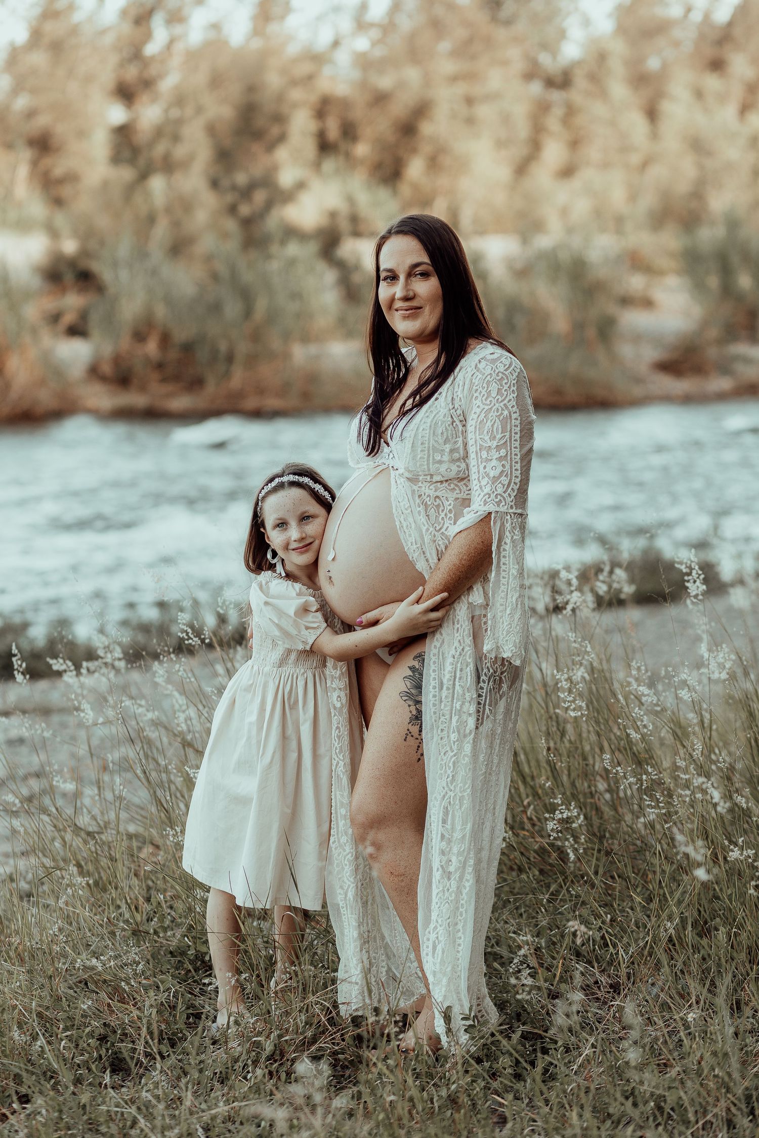 A parent and child wearing matching white lacy dresses stand together in a field during golden hour.