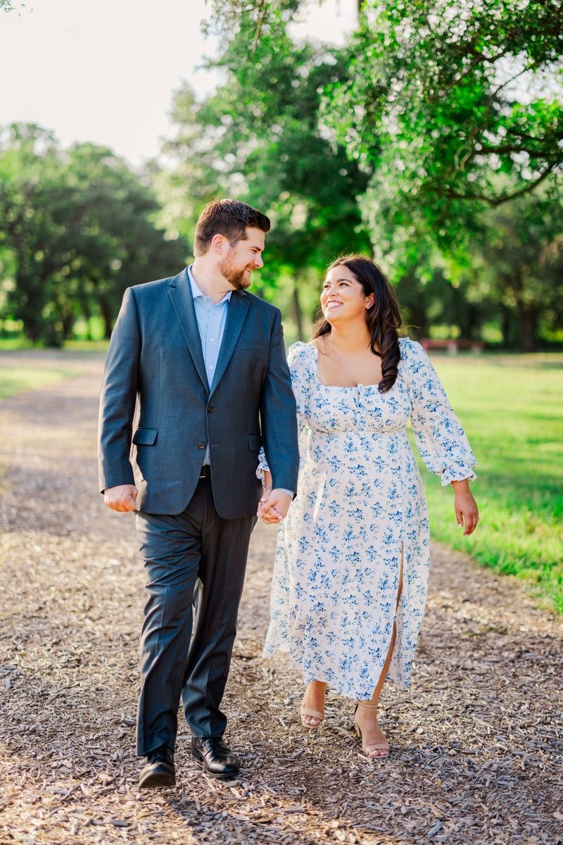 A couple walks hand-in-hand along a sunlit park path, smiling at each other in elegant semi-formal attire.