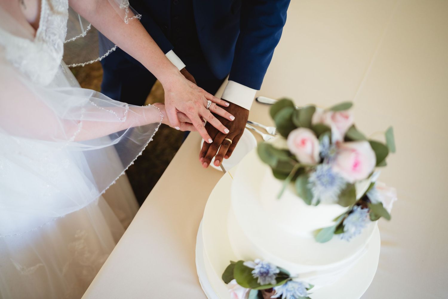 A couple's hands cut into a white wedding cake decorated with soft pink and blue flowers.