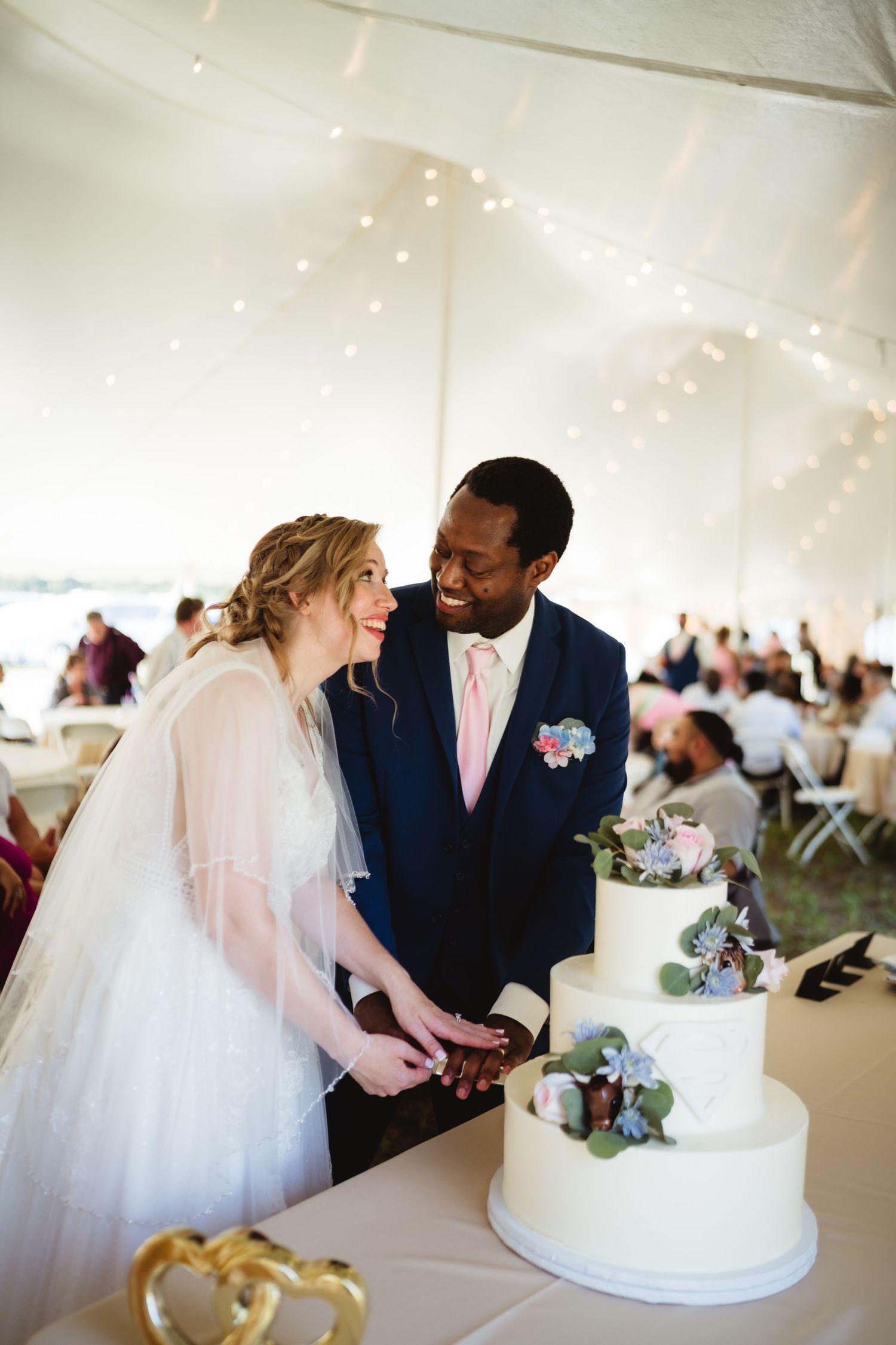 Happy newlyweds share a loving moment while cutting their elegant three-tiered white wedding cake under a tent reception.