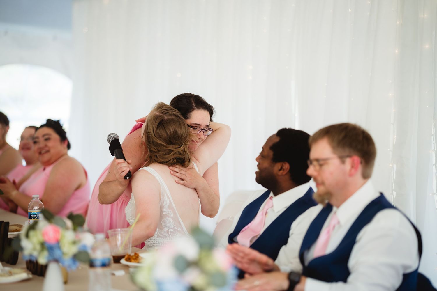 Wedding reception guests and wedding party members seated at a table with floral centerpieces during emotional moment.