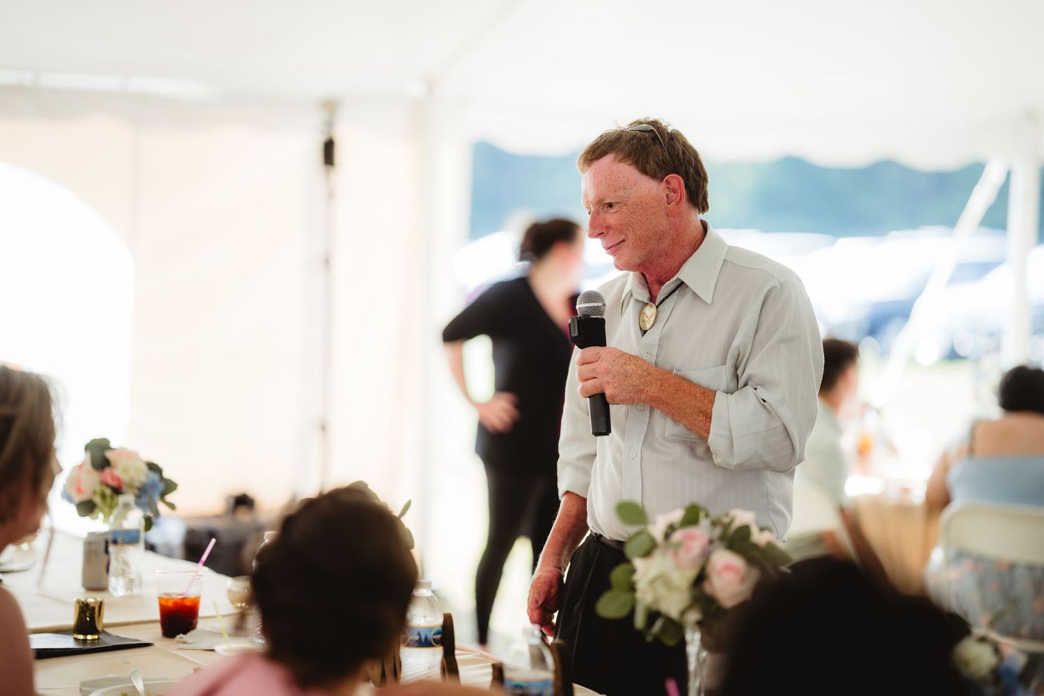 A person speaks into a microphone during a reception in a white tent with tables and floral centerpieces.