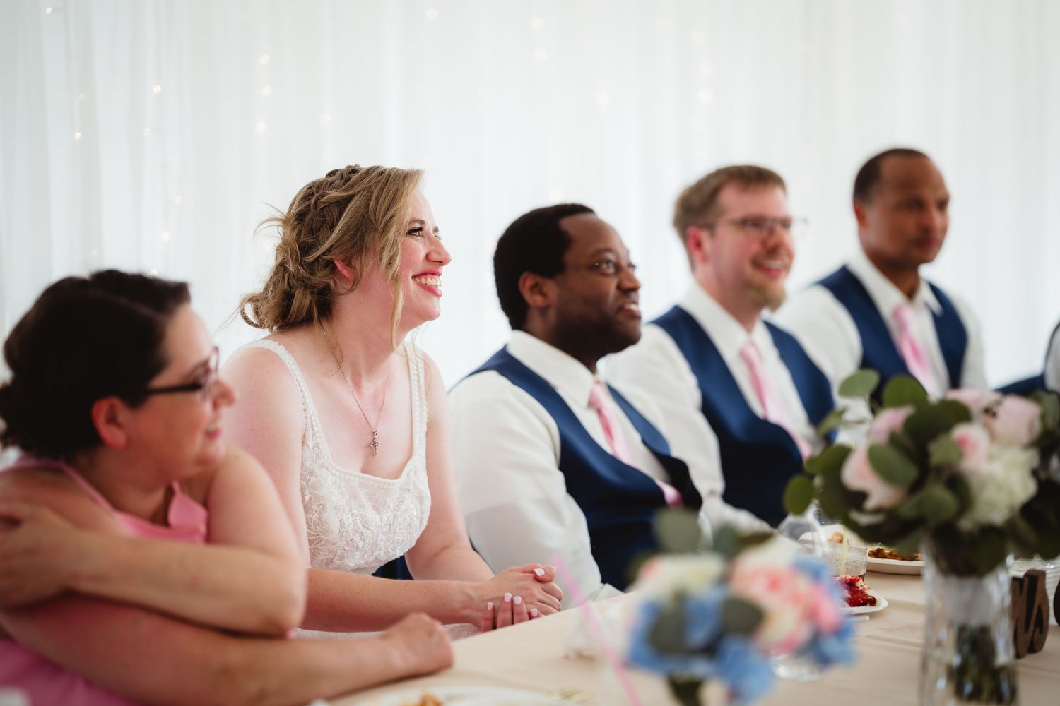 Wedding party members seated at a long table during a reception, listening intently to speeches being given.