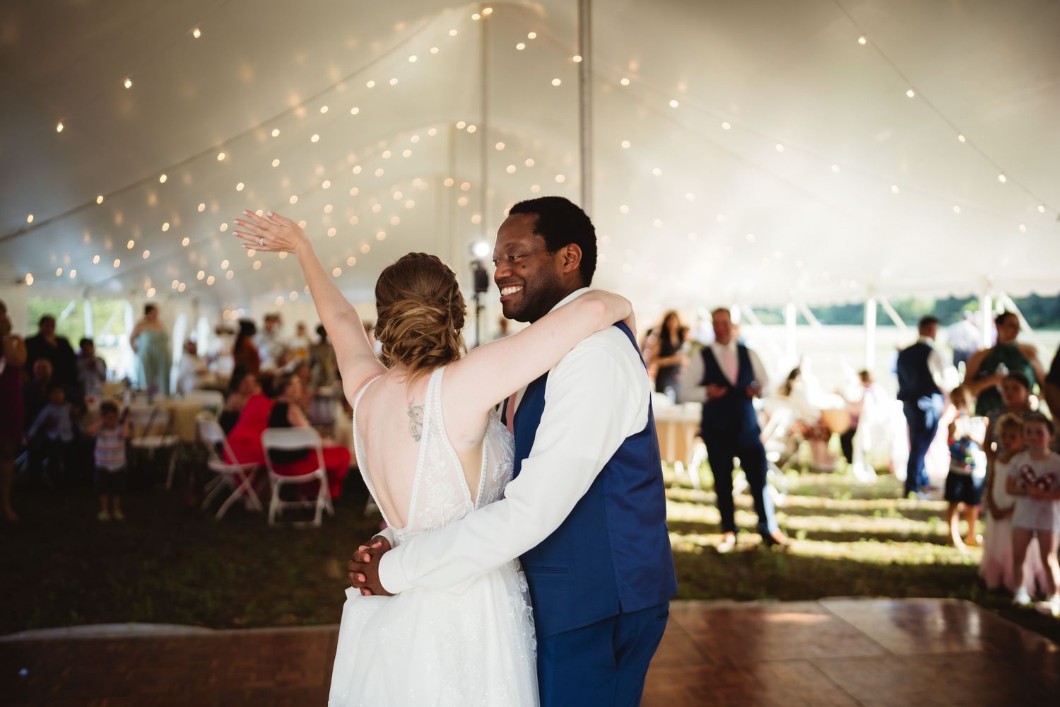 A romantic first dance at a wedding celebration under a tent with twinkling lights and guests watching in the background.