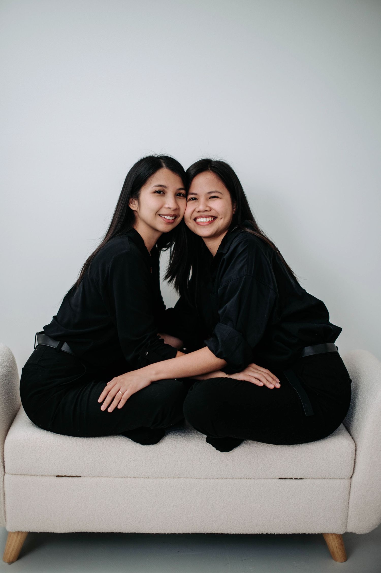 Two people wearing black clothes pose playfully together on a light colored couch in a minimalist setting.