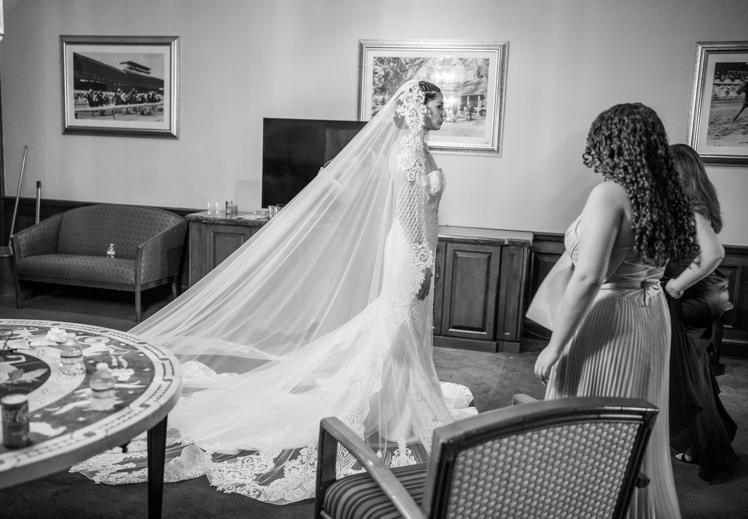 A bride in an elegant wedding gown with a flowing cathedral veil stands in an interior room with framed artwork.