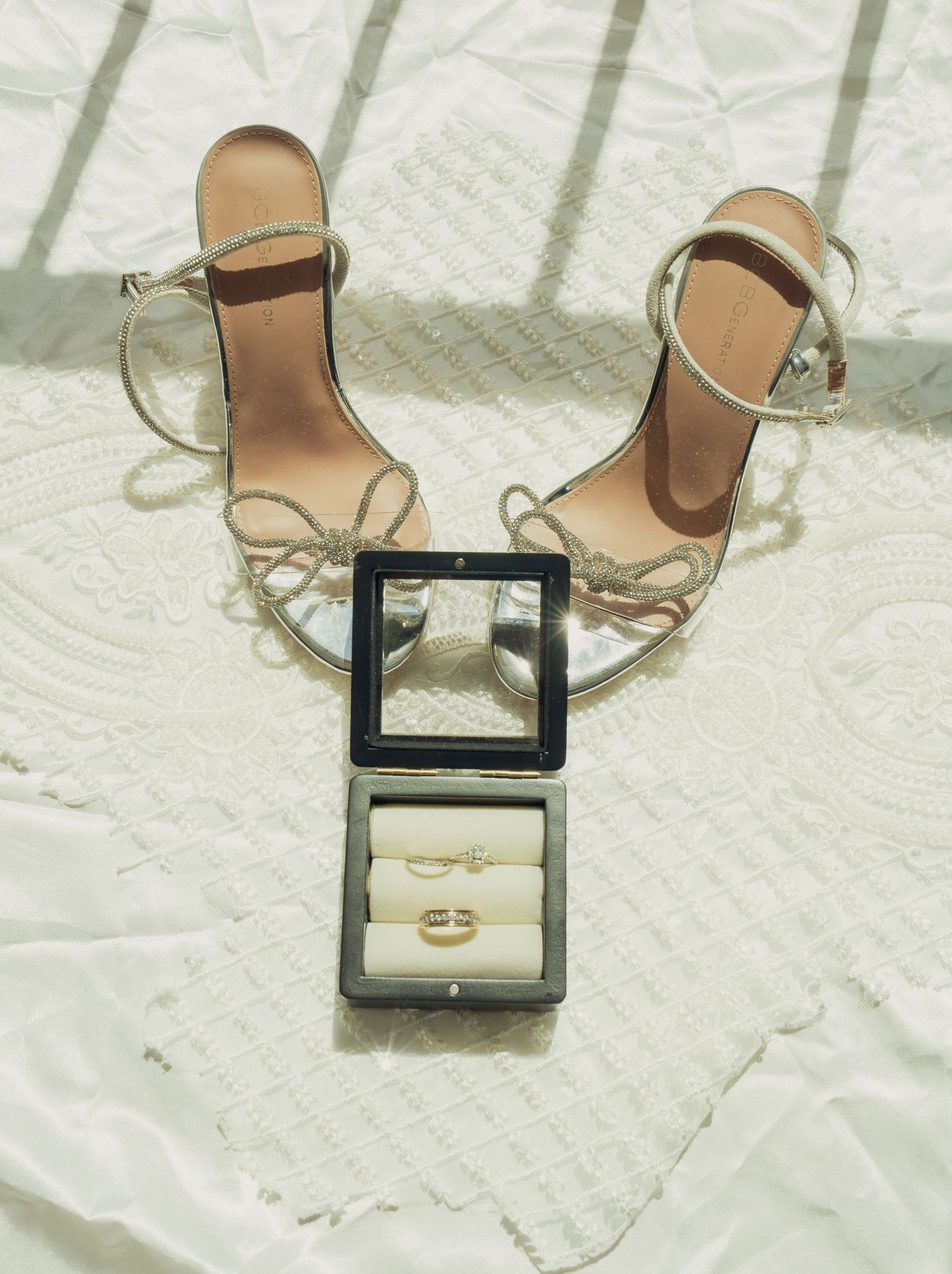 Elegant gold strappy sandals and luxury makeup items arranged on white textured fabric creating a stylish flat lay composition.