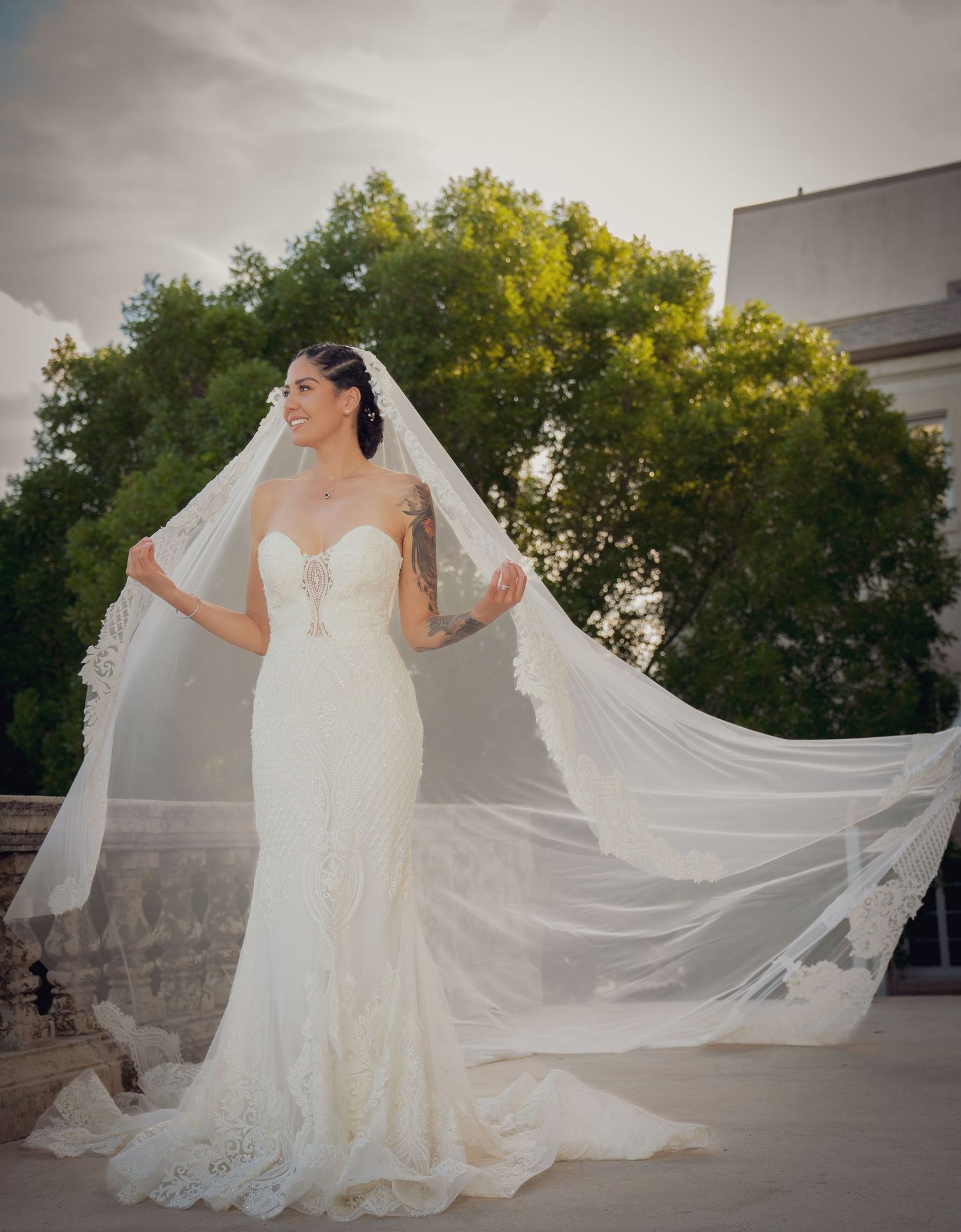 A flowing white wedding veil gracefully catches the breeze as it trails behind a bride in a lace gown outdoors.