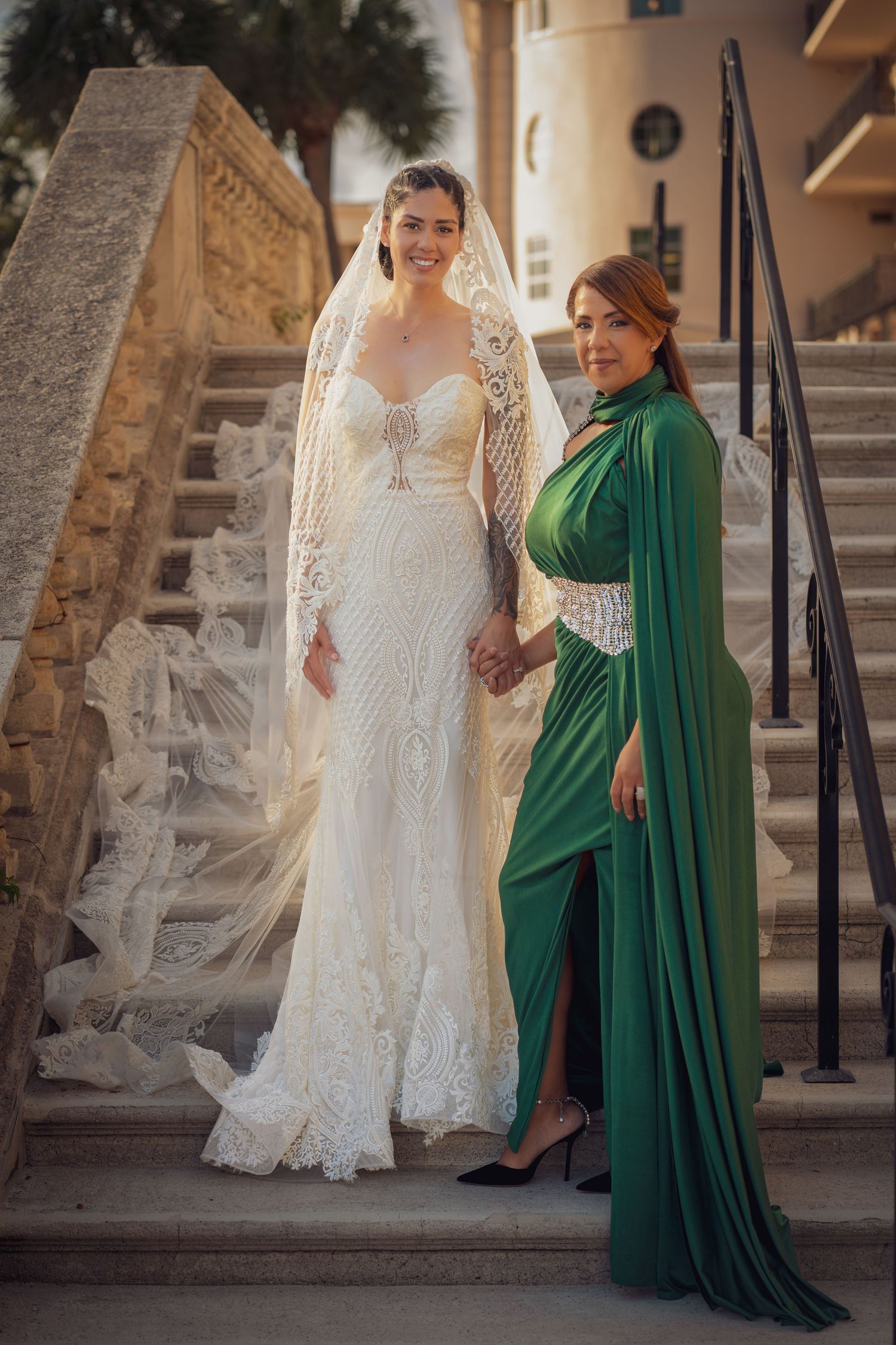 Two women pose on stone steps, one wearing an elegant lace wedding gown and veil, the other in a flowing emerald green dress.