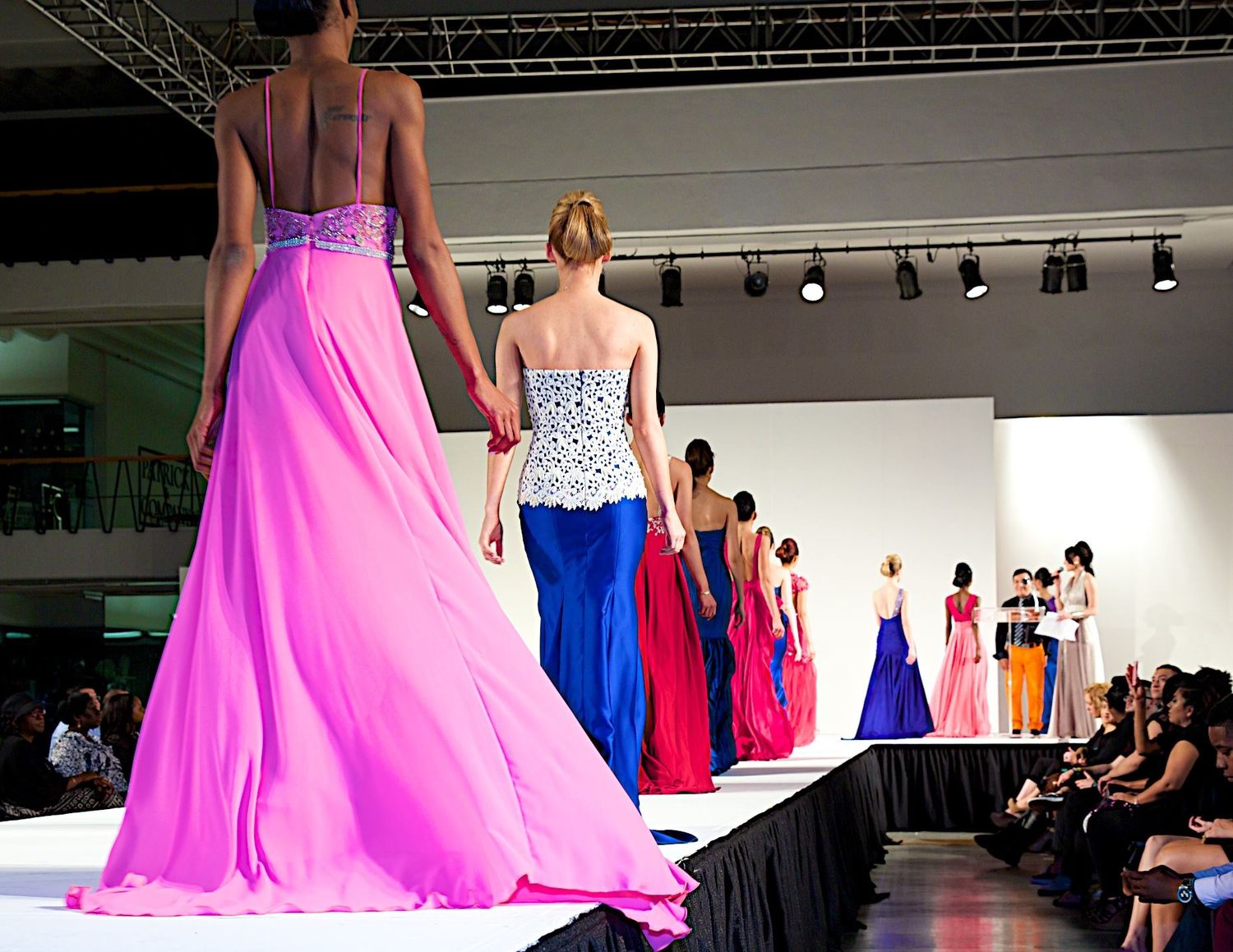 Models showcase flowing evening gowns in pink, blue and red on a runway with audience members watching the fashion show.
