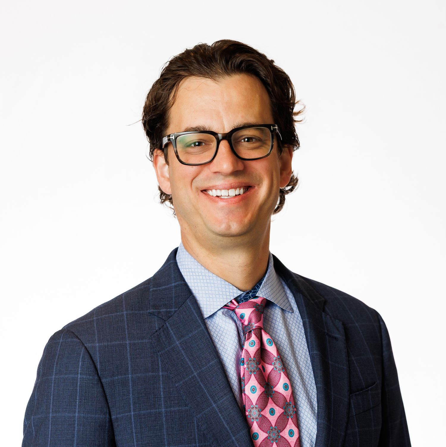 Professional headshot of person wearing glasses, navy windowpane suit jacket, striped shirt and patterned red tie.