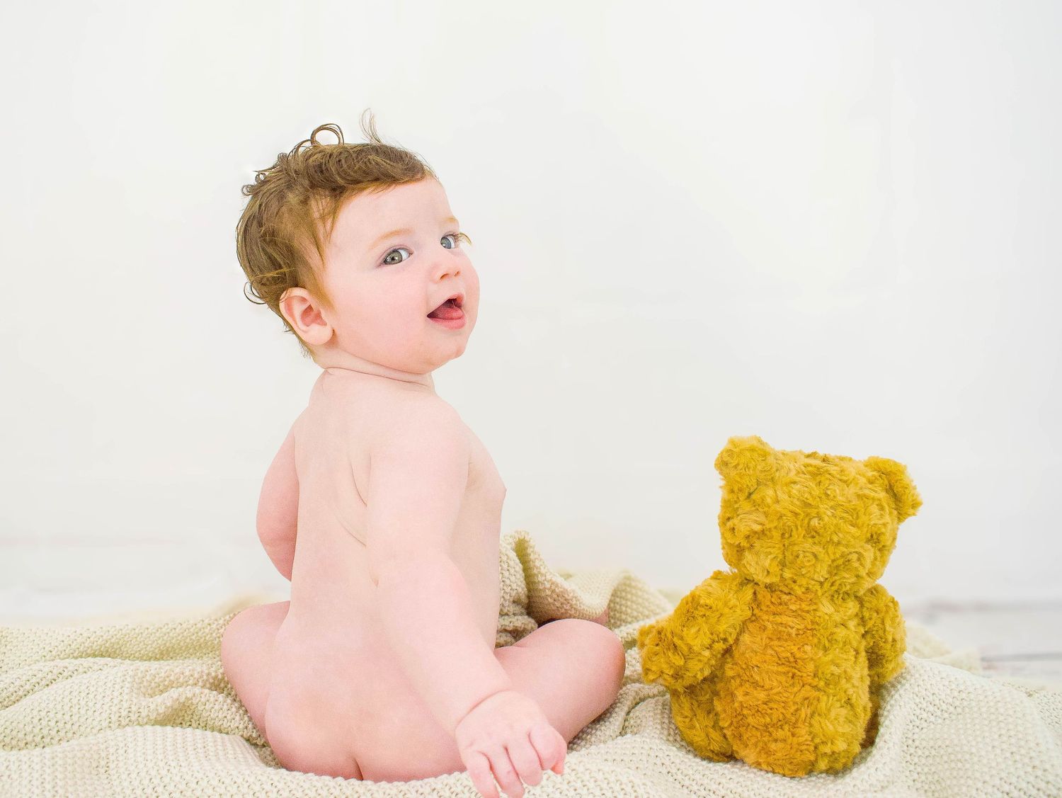Baby plays with a golden teddy bear on white blanket in sweet photo series.