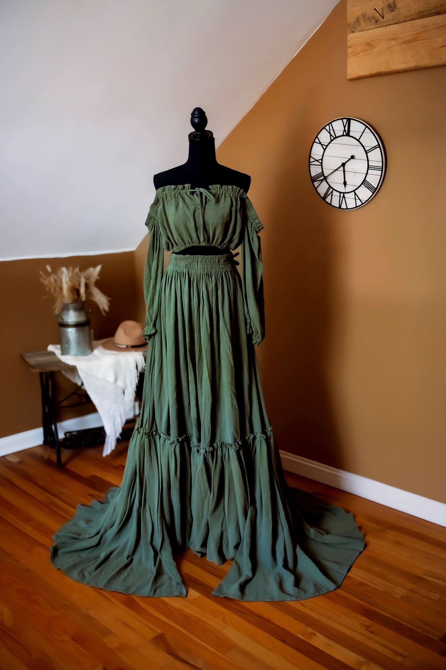A flowing champagne-colored maxi dress with bell sleeves displayed on a black mannequin against a tan wall and hardwood floor.