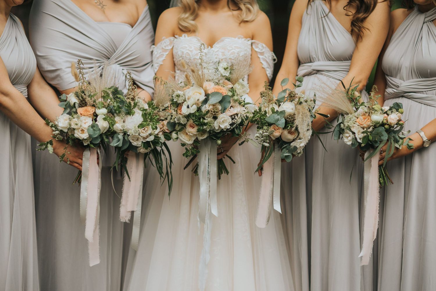 Wedding party members in light gray dresses hold elegant white and pink floral bouquets during an outdoor ceremony.