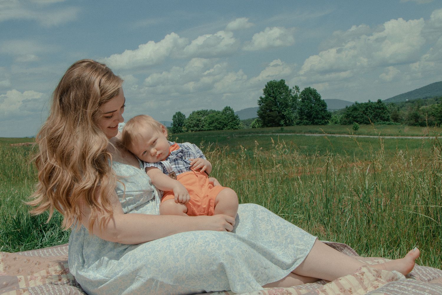 Mother sits in a sunny field holding her baby, who's resting peacefully on her chest