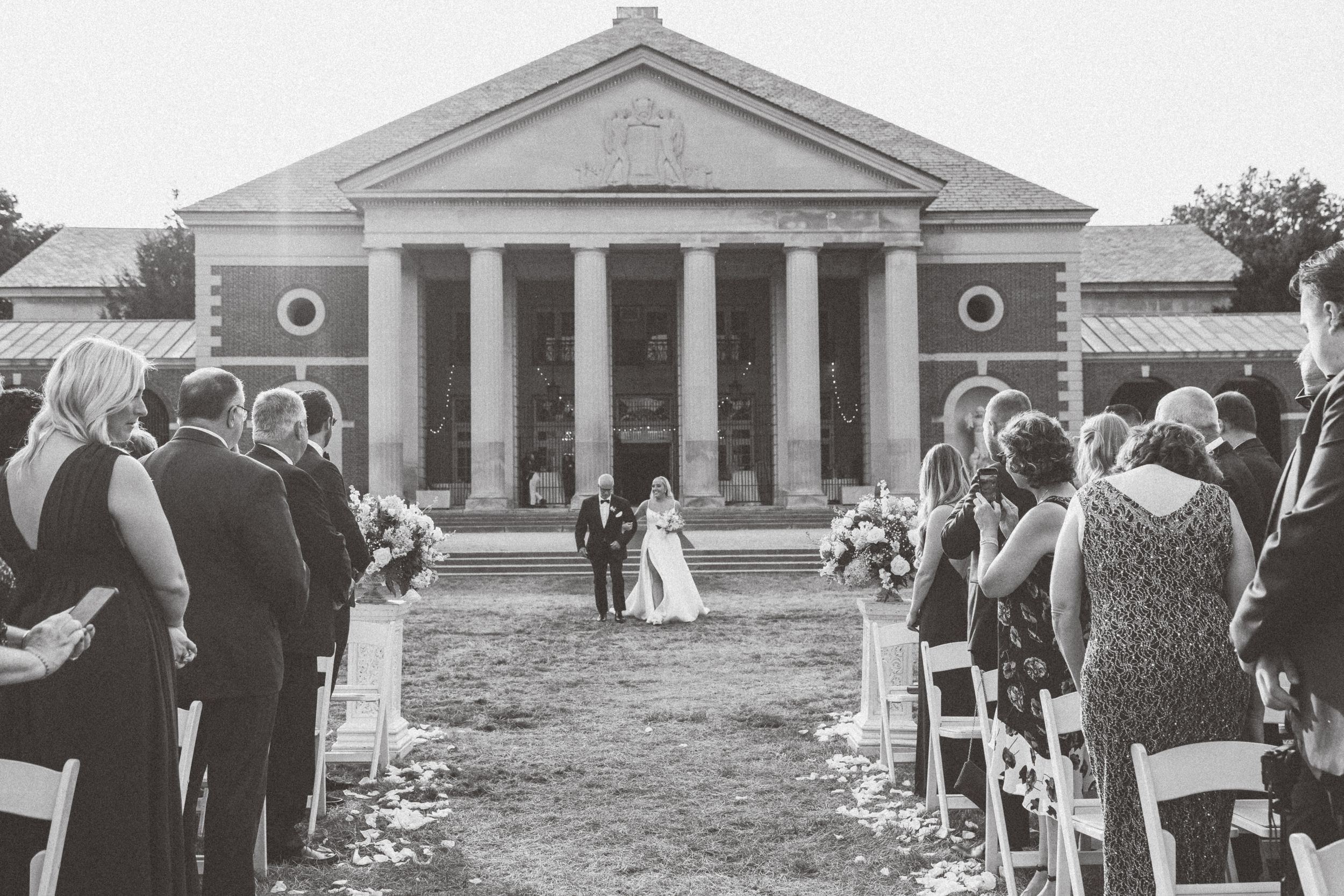 A black and white wedding ceremony taking place in front of a grand neoclassical building with columns.