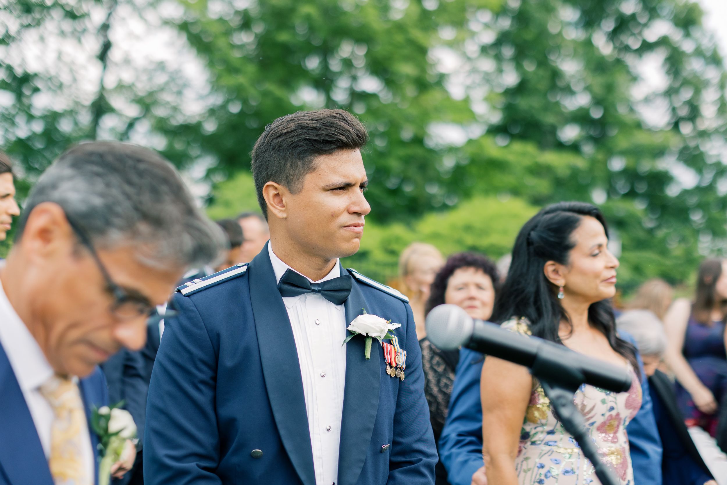 Wedding guests in formal attire watching a ceremony outdoors with trees in the background.