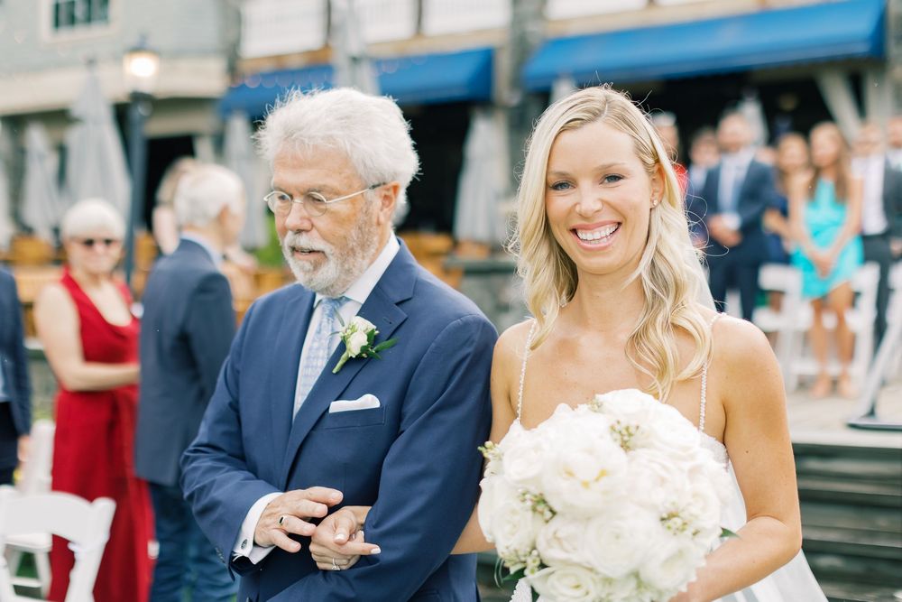A smiling pair walking arm in arm at an outdoor wedding ceremony with a white peony bouquet.