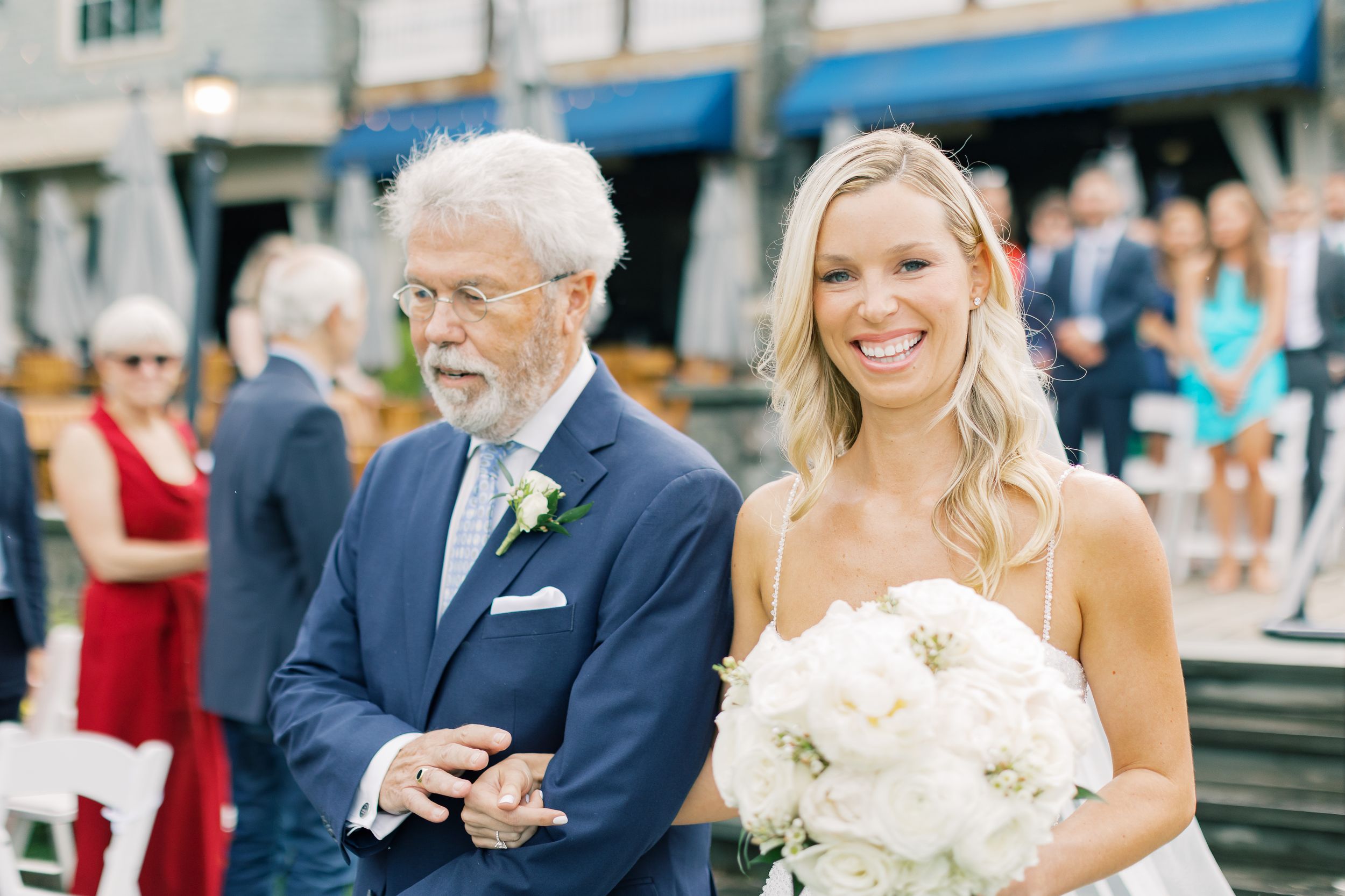 A smiling pair walking arm in arm at an outdoor wedding ceremony with a white peony bouquet.